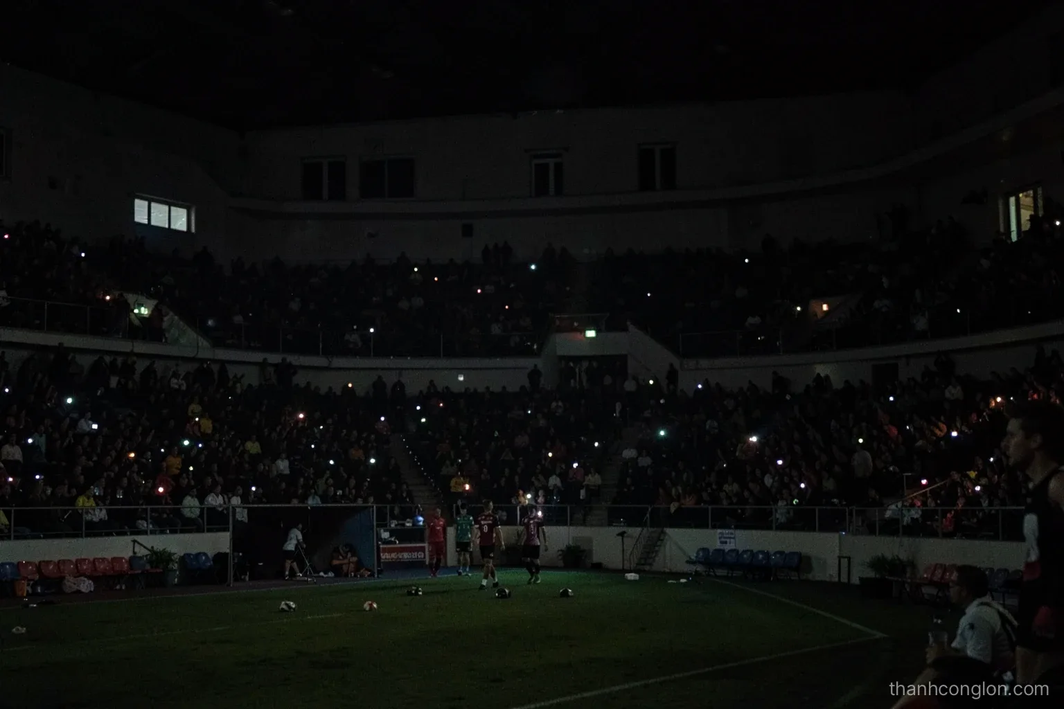 Darkened stadium seen from the upper stands during a power outage, scattered phone lights glowing among the crowd, players standing on the dim field below