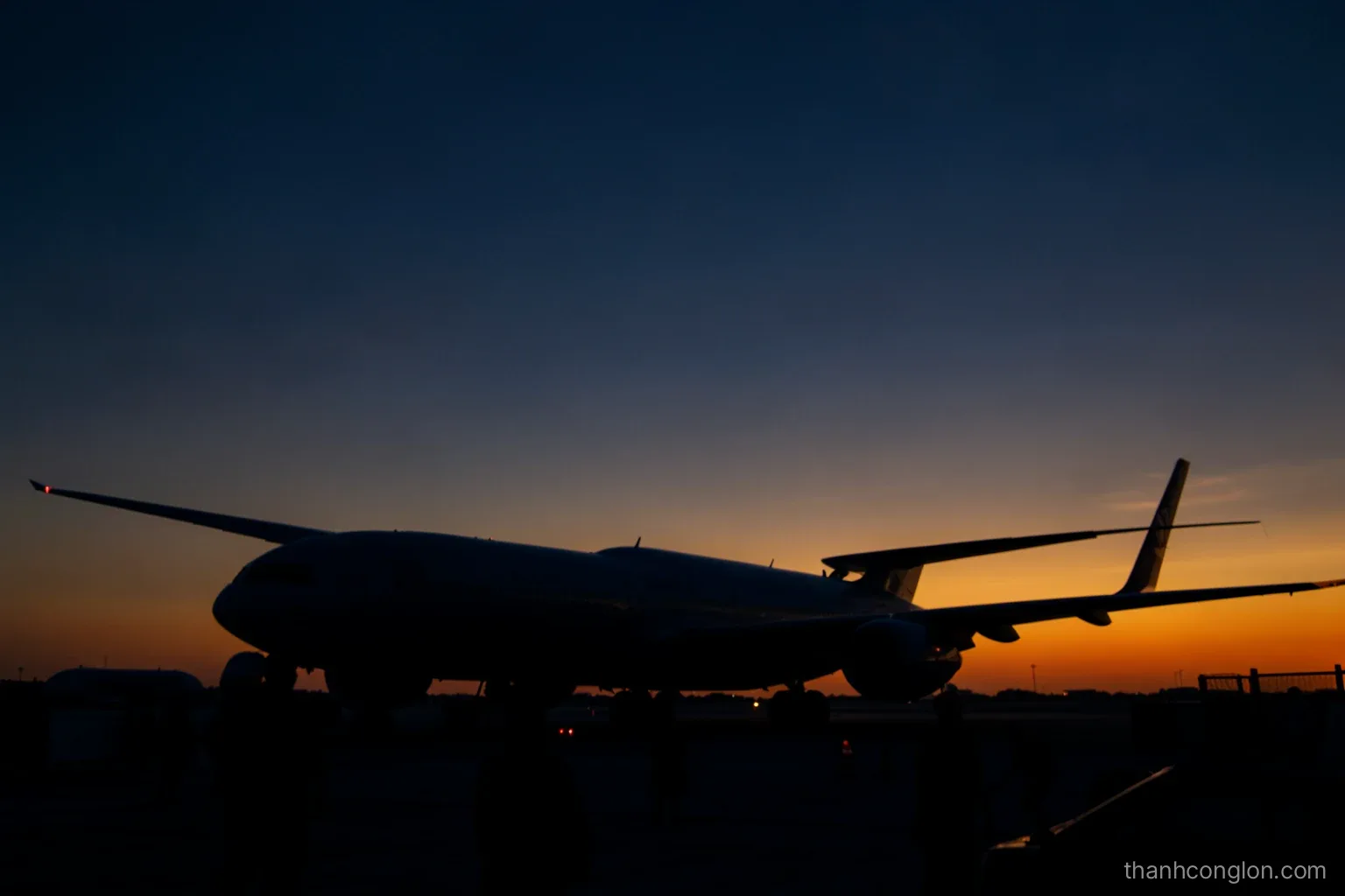 Silhouette of a large passenger jet parked at a quiet airport gate before sunrise, ground crew figures in the foreground seen from behind, deep navy and amber sky